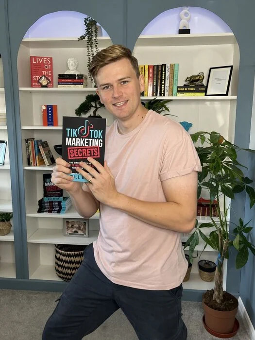 Will Bower holding a copy of his book, TikTok Marketing Secrets, in front of a bookshelf.