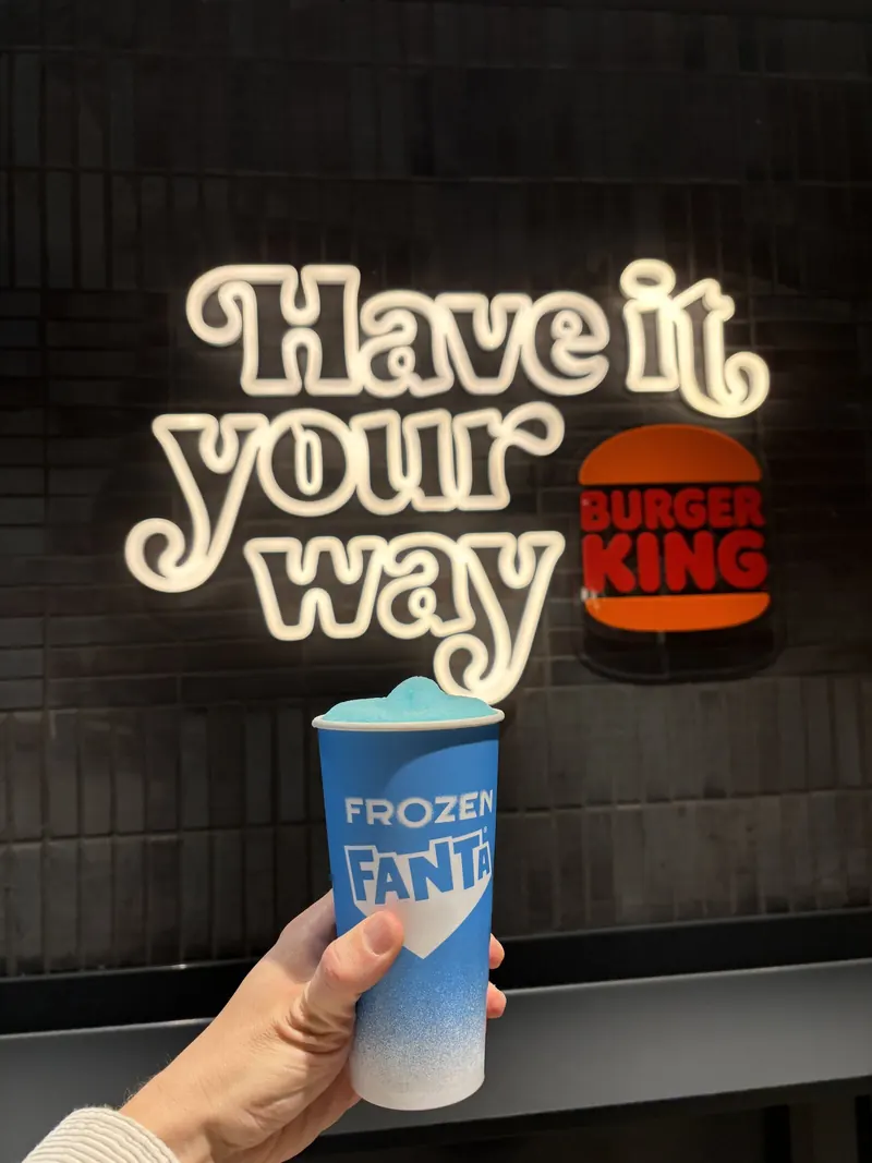 A Fanta Frozen cup held up in front of a neon Have it your way sign.