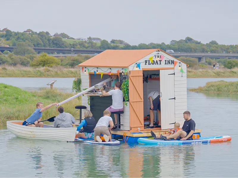 The Float Inn: a small pub with a peach roof and bunting, floating on the water with family on paddleboards and a rowing boat beside it.