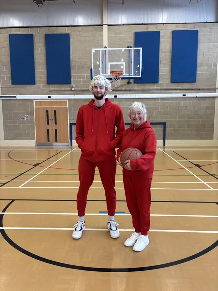 The stunt double and the granny in matching red tracksuits on a sports hall court, the granny holding a basketball.