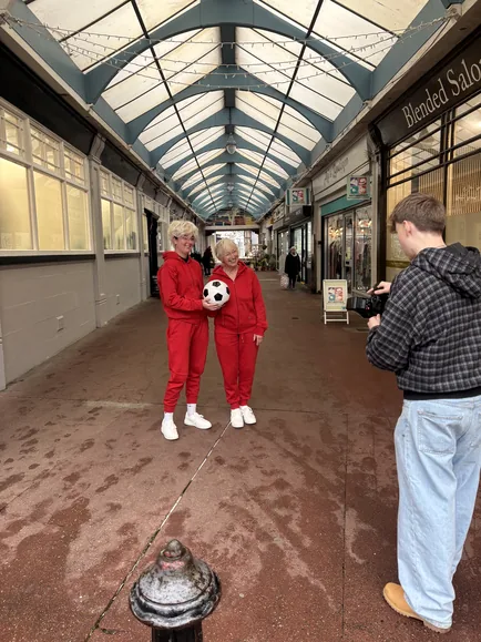 The stunt double and the granny with a football in a covered arcade, filmed by a camera operator.