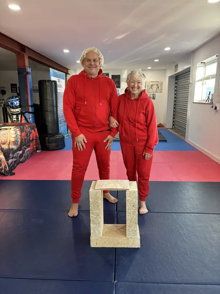 The stunt double and the granny in matching red tracksuits on a dojo mat, with the wood-chop board on blocks.