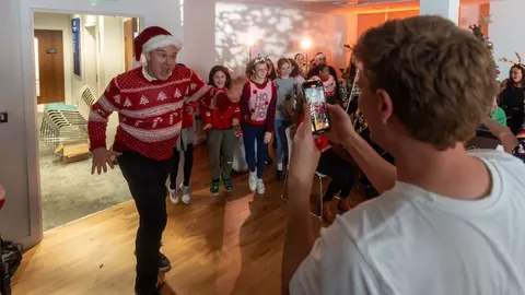 Ed Davey in a Christmas jumper dancing down a corridor with a children's choir, while Will films on a phone.