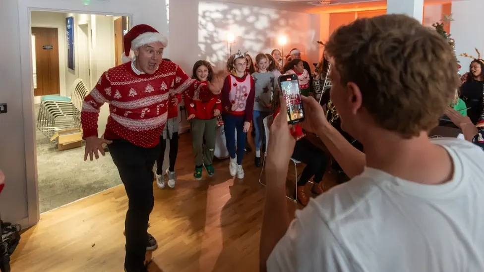 Ed Davey in a Christmas jumper dancing down a corridor with a children's choir, while Will films on a phone.