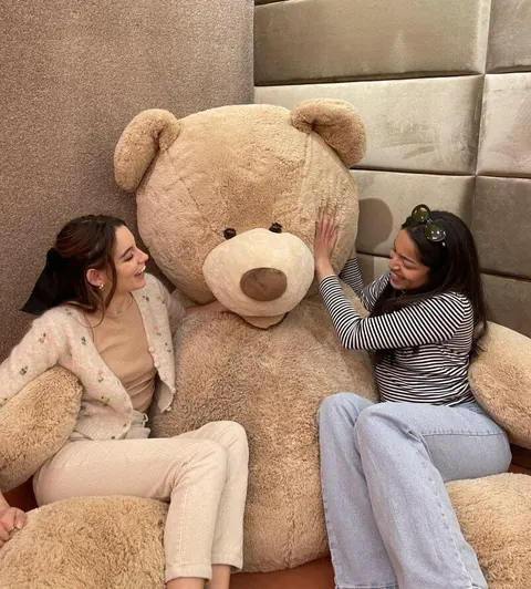 Two women on a bed beside a giant teddy bear in the MAC Village Velvet Teddy bedroom.