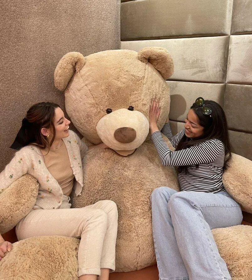 Two women on a bed beside a giant teddy bear in the MAC Village Velvet Teddy bedroom.