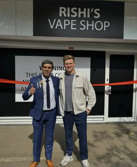 Will with a Rishi Sunak lookalike in a suit outside a shop signed Rishi's Vape Shop, with a ribbon across the door.