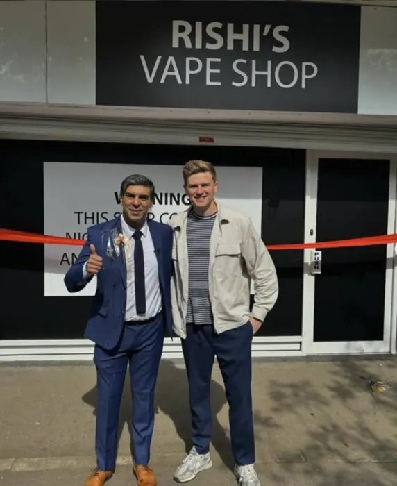 Will with a Rishi Sunak lookalike in a suit outside a shop signed Rishi's Vape Shop, with a ribbon across the door.