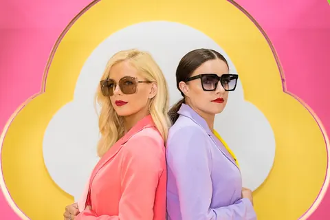 Two women in sunglasses and pastel suits, back to back, in front of a yellow and pink cloud set.