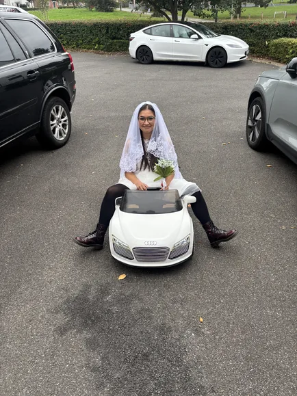 The character in a wedding dress and veil, sitting in a white toy car in a car park.